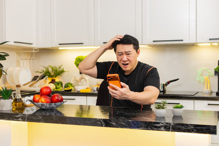 Asian man in home kitchen looks at orange smartphone with shocked expression after online game loss and lottery failure. Bad news from casino app brings stress and frustration about money and result.の写真素材