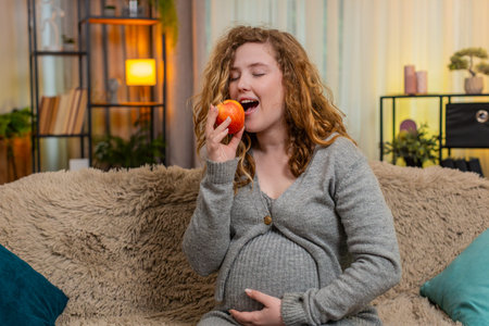 Pregnant young happy woman sits on home sofa, gently touching belly with one hand, taking a bite of fresh apple, enjoying nutritious snack. Girl feels peace and satisfaction, embracing healthy choicesの写真素材