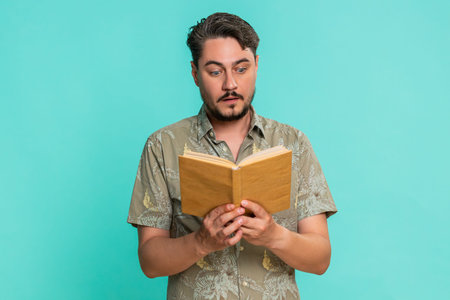 Middle-aged man reads book with focused expression, raised eyebrows, reacting to intriguing turn in plot or emotional moment. Young adult guy isolated on blue background engaged in reading literature.の写真素材