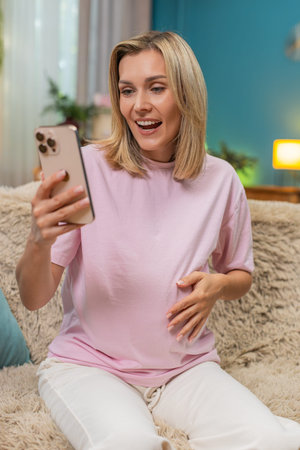 Pregnant middle-aged woman at home smiles typing on smartphone celebrating good news message and lifting victory win hand sign. Happy Caucasian girl on sofa feels anticipation texting on mobile phone.の写真素材