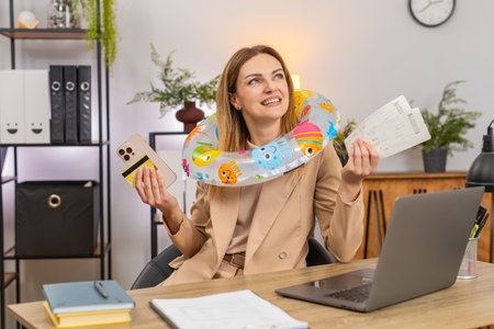 Middle-aged businesswoman at home office holds credit card with smartphone pays tickets smiles ends work on laptop. Freelancer girl at table puts swim ring on head packs list plans beach vacation tripの写真素材