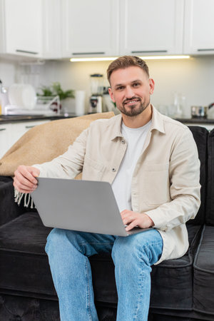 Young man at home opens laptop starts work launches system and types fast with focus and confident smile appears. Caucasian guy on sofa checks tasks writes notes shows productive mood with clear goal.の写真素材
