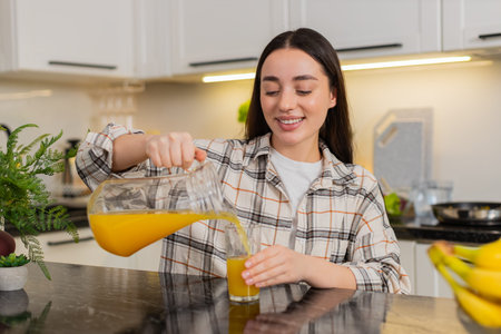 Young woman pours fresh orange juice from a glass pitcher into a tumbler in home kitchen. Natural citrus beverage supports hydration wellness, morning routine. Cheerful girl chooses healthy lifestyle.の写真素材