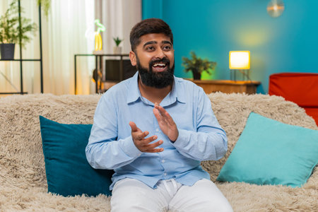 Indian man at home claps hands after online game win smiles bright feels happy celebrates loudly. Arabian guy on sofa applauds big news huge joy moment shows victory gesture great energy celebrationの写真素材