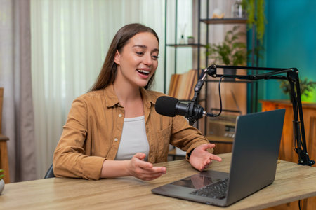 Young woman at home table speaks into studio microphone during live stream sharing practical tips,...