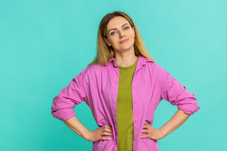 Middle-aged woman places hands on hips in firm confident pose, expressing strength, determination, leadership. Young adult girl isolated on blue background shows assertive stance and empowered mindsetの写真素材