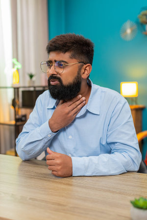 Man at home clutches sore throat struggles to swallow feeling sharp pain infection virus fever. Guy at table looks sick contorts face griping pain seeks relief medicine worrying healthの写真素材