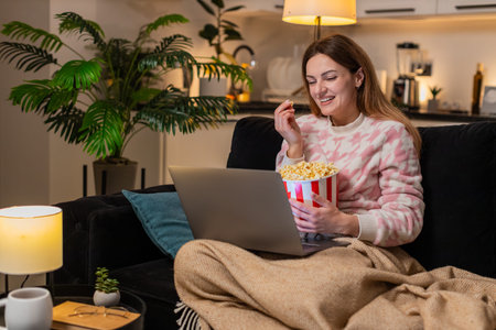 Middle-aged woman at night home with laptop and popcorn watches trailer or film online laughs comments. Caucasian girl on sofa at evening enjoys comedy reacts to funny moments talks to screen smiles.の写真素材