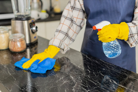 Woman sprays cleaner on countertop wiping surface with microfiber cloth wearing gloves for...