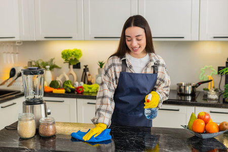 Young woman wipes kitchen table with cloth and cleaning spray wearing gloves for home hygiene and...