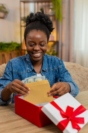 African American young woman sitting at table on home sofa happily checks envelope full of cash...