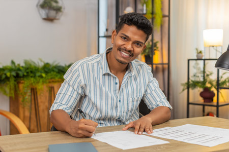 Happy man at home finds contract reads terms and signs with pen smiles with confidence. Guy at table...