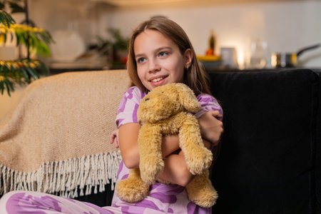 Teenager girl at night home holds plush puppy toy near face smiles soft cuddle gentle mood. Caucasian child kid on sofa at evening hugs toy close feels safe calm joy slow breath warm heart sweet care.の写真素材