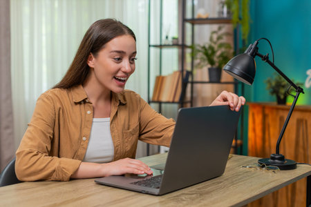 Young woman sitting at home table opens laptop sees project results smiling widely celebrating...