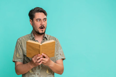 Middle-aged man reads book with focused expression, raised eyebrows, reacting to intriguing turn in plot or emotional moment. Young adult guy isolated on blue background engaged in reading literature.の写真素材