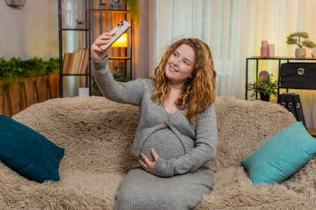 Pregnant young woman takes a selfie on smartphone, gently touching her belly, smiling with warmth and love for future baby. Girl captures special moments, celebrating connection and motherhood at homeの写真素材