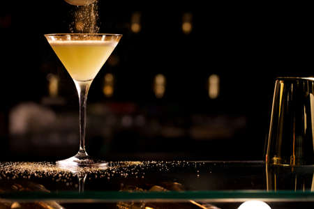 dropping sugar onto a ginger cocktail, yellow cocktail ob a table, on dark background. Bartender with glass and sugar preparing cocktail at bar.の写真素材