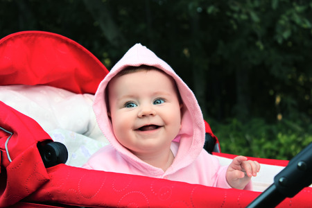 Happy little girl dressed in pink is sitting in the red pram, looking around and smiling during  a stroll in the park. Trees on the background.の写真素材