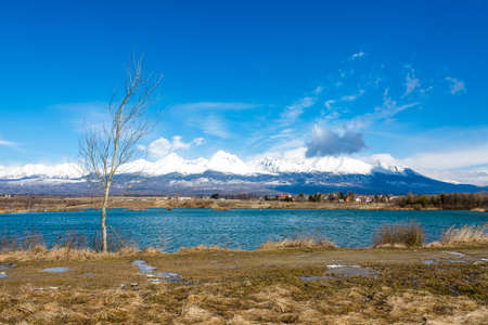 Slovakia: Big Tatras (Vysoke Tatry) on the spring. Big mountains with the snow and clouds. The lake in foreground. Small village near the mountain and hillの写真素材