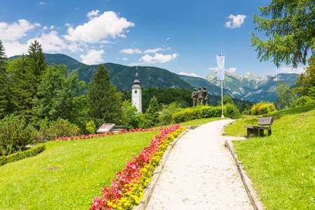 View of Bohinj church in Ribcev Laz village, from the park with statue. Triglav and mountains in the background, park with grass and flowers in foreground.の写真素材