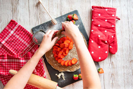 Galette with strawberries preparation, detail of hands of cooker. Pie cake placed on black desk and wooden table with cooking tools on the side.の写真素材