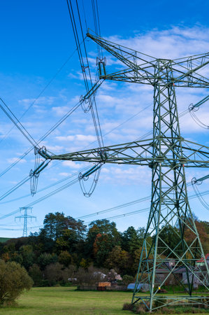 A landscape photo of power lines and transmission towers stretching across an open field with a scenic backdrop of hills and a partly cloudy sky. The image captures the intersection of nature and industry, highlighting the role of infrastructure in rural landscapesの写真素材