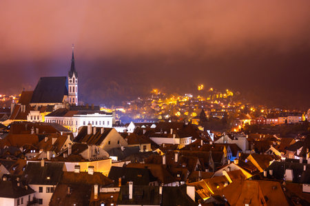 A panoramic night view of Cesky Krumlov, showcasing the rooftops, illuminated buildings, and the Church of St. Vitus beautifully lit up against the dark skyの写真素材