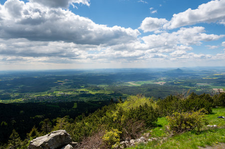 Panoramic view of green fields and forested hills near Jested, with the sharp peak of Ralsko mountain visible in the distance under a blue sky with scattered clouds.の写真素材