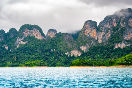 View of Khao Sok Lake in Krabi province, Thailand, with towering limestone cliffs covered in tropical rainforest rising above the calm surface of the lake.の写真素材
