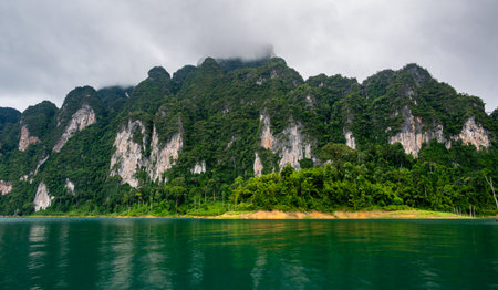 View of Khao Sok Lake in Krabi province, Thailand, with towering limestone cliffs covered in tropical rainforest rising above the calm surface of the lake.の写真素材