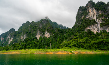 View of Khao Sok Lake in Krabi province, Thailand, with towering limestone cliffs covered in tropical rainforest rising above the calm surface of the lake.の写真素材