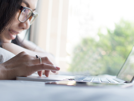 Asian girl is typing and talking with a other person with her laptop on a bed in her vacation at her house.の写真素材