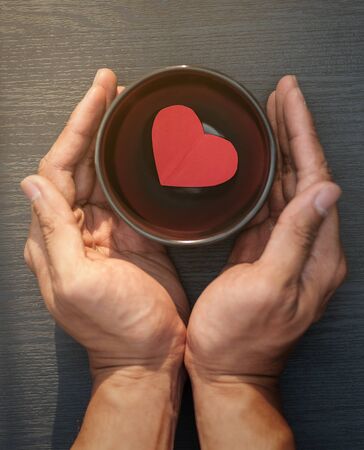Two hands protect a  red paper heart in a black bowl  from the sunlight on wooden panel.の写真素材