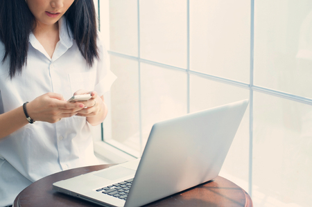 Asian girl is holding a mobile phone and a laptop on round wooden table at her room on white background.の写真素材