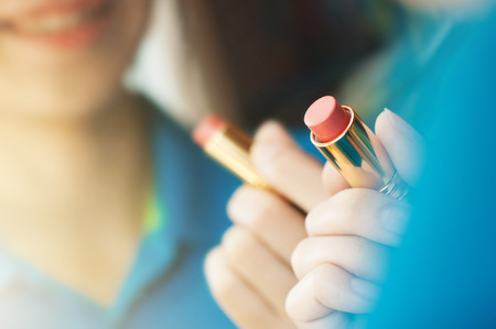 Asian woman is applying makeup and looking in the mirror. Closeup of lipstick. Hand of woman is holding red lipstick. Woman applying makeup and smiling.の写真素材