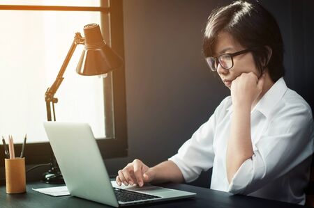 Close up of asian girl thinking about her work. Eyeglasses girl looking and typing her laptop with sunlight from window. Business woman.の写真素材