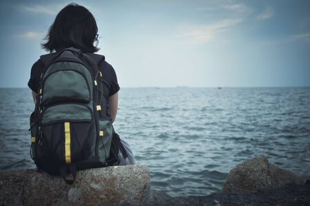 Woman traveller sitting on rock for resting and looking to sea with cloudy sky. Copy space. Rear viewの写真素材