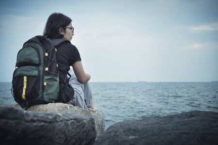 Woman traveller sitting on rock for resting and looking to sea with cloudy sky. Copy space. Rear viewの写真素材
