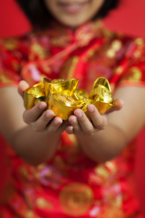 Chinese new year. Asian glasses woman with cheongsam holding gold Ingots on red background. Girl is smiling.の写真素材