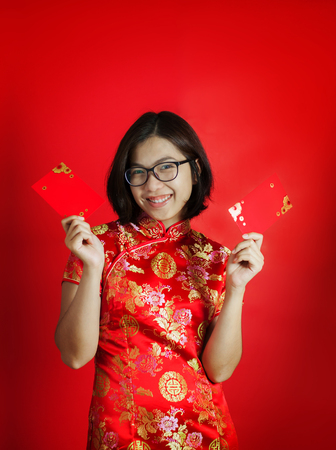 Chinese new year. Asian glasses woman with cheongsam holding red envelopes on red background. Girl is smiling.の写真素材