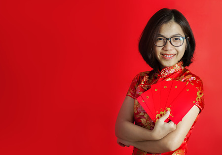 Chinese new year. Asian glasses woman with cheongsam holding red envelopes on red background. Girl is smiling.の写真素材
