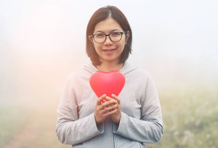 Valentine's concept. Asian woman holding red heart on fog and grass field background. Copy space. Vintage or retro color. winter.の写真素材