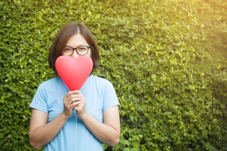 Valentines concept. Asian glasses woman holding red heart on green grass background. Copy space.の写真素材