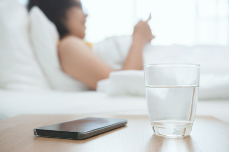 Close up Woman  glass of water with smartphone on foreground. Beautiful Asian girl reading her book and lying on bed.の写真素材