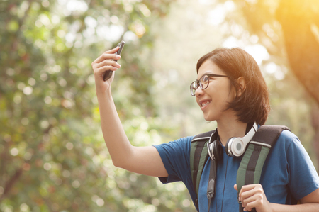 Asian glasses woman hiker with backpack taking nature photo with her cell phone. Beautiful girl smiling on natural background.の写真素材