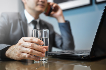 Close up Asian Businessman in grey suit holding glass of water. Man using mobile phone with laptop on foreground.の写真素材