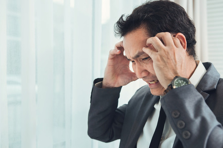 Asian businessman has headache from migraine from overworked. Illness, exhausted, disease, tired concept.の写真素材