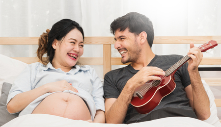 Beautiful pregnant woman and handsome husband are smiling and playing Ukulele or acoustic guitar on bed.の写真素材