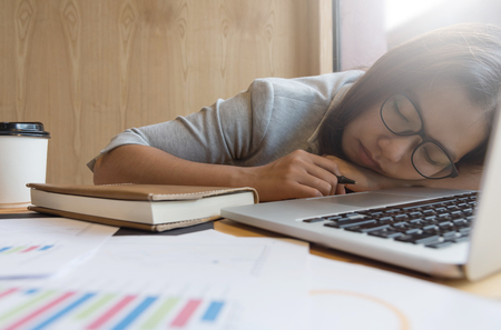 Tired Asian business woman sleeping from overwork with laptop, Graph financial diagram documents and a cup of coffee at the office.の写真素材