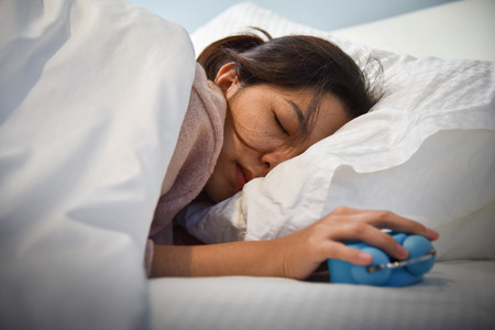 Sleepy Asian woman extending hand to Alarm clock on her bed. Female having trouble waking up. Stress from overtime working concept. Nightmare.の写真素材